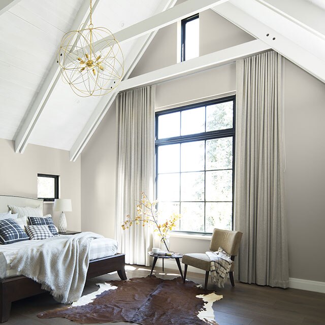 A sunlit bedroom with light gray-painted walls, a white cathedral ceiling with beams and a large paned window.
