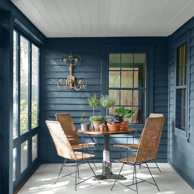 Patio couvert bleu au plafond en planches à feuillure blanches avec plancher blanc, table ronde, plantes en pot et chaises en osier.