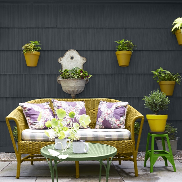 Patio chaleureux avec canapé rembourré en osier jaune-vert et coussins colorés ainsi que deux tables vertes devant un parement noir clair sur lequel sont accrochées des plantes en pot.