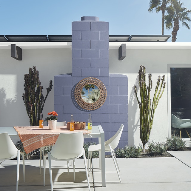 Voici une maison en stucco blanc dont la cheminée extérieure lilas flanquée de deux cactus agrémente le coin à manger au mobilier blanc de ce patio.
