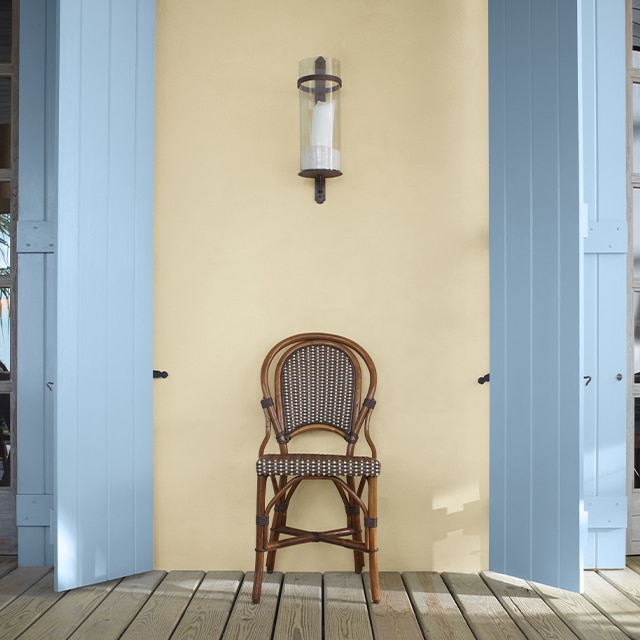 Un porche arborant une chaise entre deux volets peints en bleu clair sur un mur peint en jaune pâle.