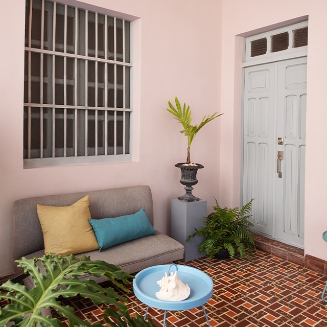 Une jolie galerie qui fait office de boudoir présente des murs rosés, une porte grise, un petit sofa, une table bleue, des plantes tropicales et un flamboyant plancher à carreaux rouges. 