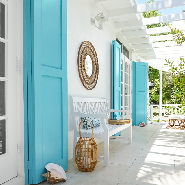 Galerie blanche rappelant les tropiques avec pergola fixée à la maison, longs volets turquoise, banc blanc et accessoires en osier.